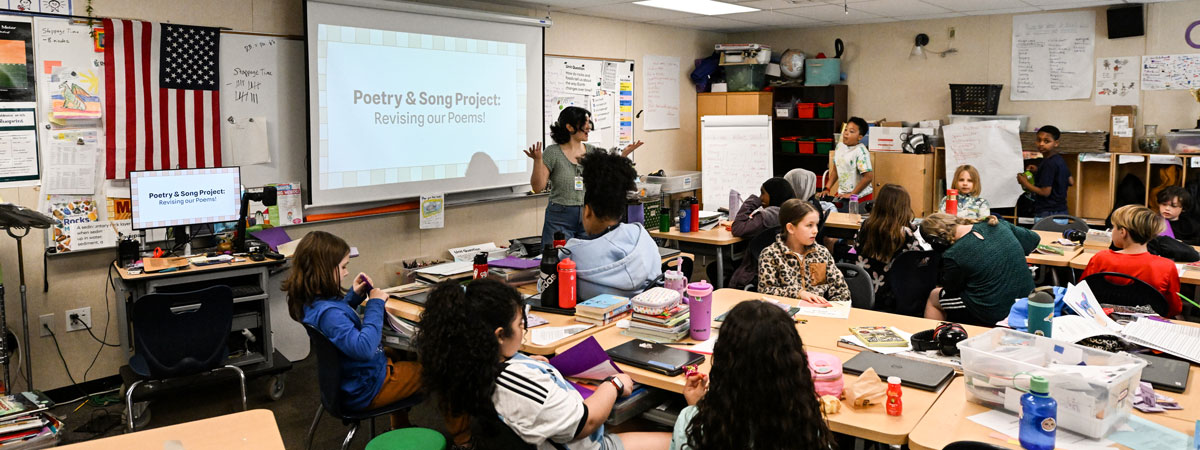 Gabriella in a classroom with 4th graders. Text projected on a screen behind her reads Poetry and Song Project: Revising our poems!