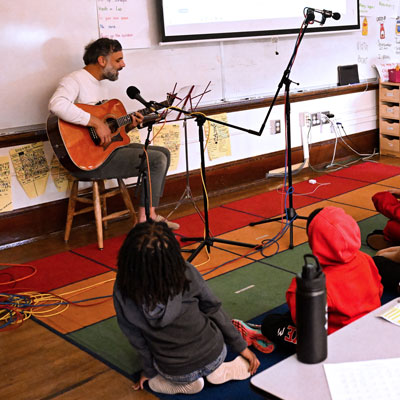 Tito sits playing acoustic guitar at a microphone in an elementary classroom. Two students sit on the floor in the foreground.