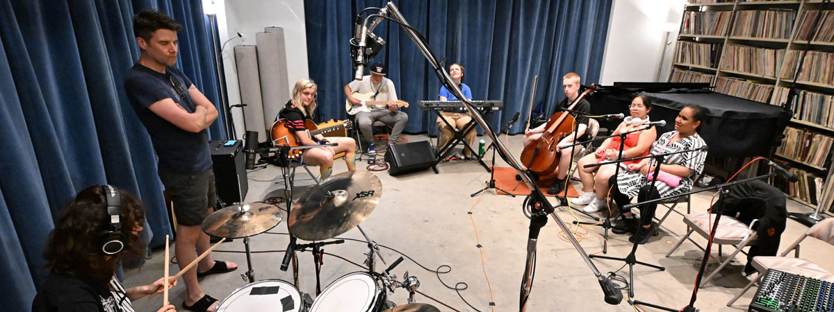 Eight people in a recording studio. One at a drumkit, one standing, two with guitars, one at a keyboard, one cello, and two at microphones.