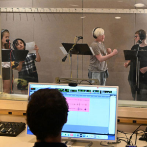 Four people in a recording studio wearing headphones at microphones. In the foreground, someone at a computer with an audio editing program.