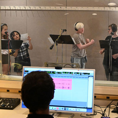 Four people in a recording studio wearing headphones at microphones. In the foreground, someone at a computer with an audio editing program.