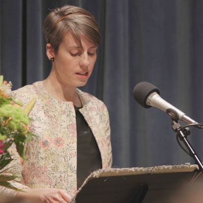 Elissa stands at a microphone, looking down. Flowers on the left edge of the frame.
