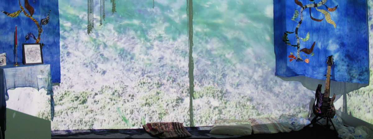 Gallery space with objects on a pedestal, a guitar, and hanging tapestries. Ocean images projected on the white wall.