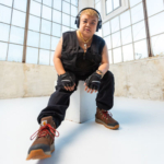 Nic seated in a white room with a grid of glass windows in the back ground. They wear headphones, black clothes, fingerless gloves, and boots.