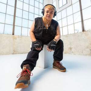 Nic seated in a white room with a grid of glass windows in the back ground. They wear headphones, black clothes, fingerless gloves, and boots.