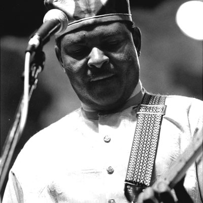 Black and white photo of Frank Ulwenya playing electric guitar with a microphone in front of him.
