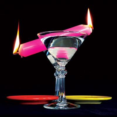 Two martini glasses with red and yellow plates on either side, with a pink candle lighted on both ends suspended between them.